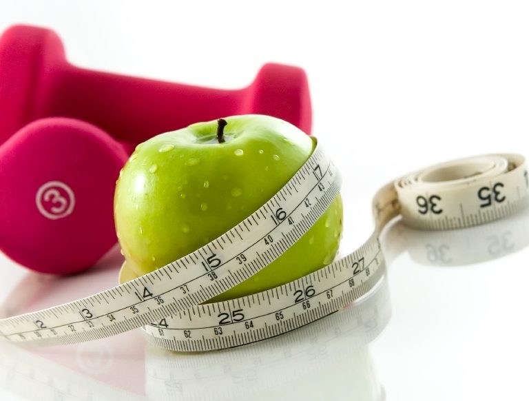 Diet and exercise Fresh appetizing apple and brightly colored dumbbells tied with a measuring tape. Slight reflection, white background, focus on the apple